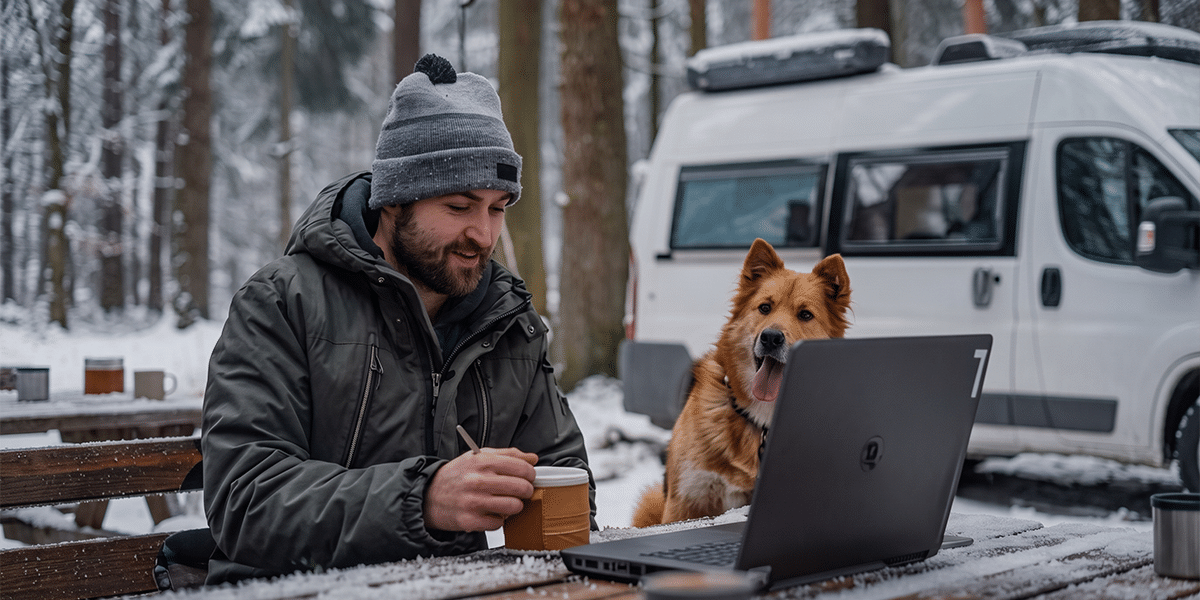 Man working on a laptop at a snowy campsite picnic table with his dog and camper van behind him